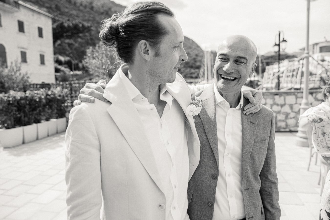 Two men smiling and embracing outdoors at a formal event; one wears a light suit with a boutonniere, and the other a gray suit.