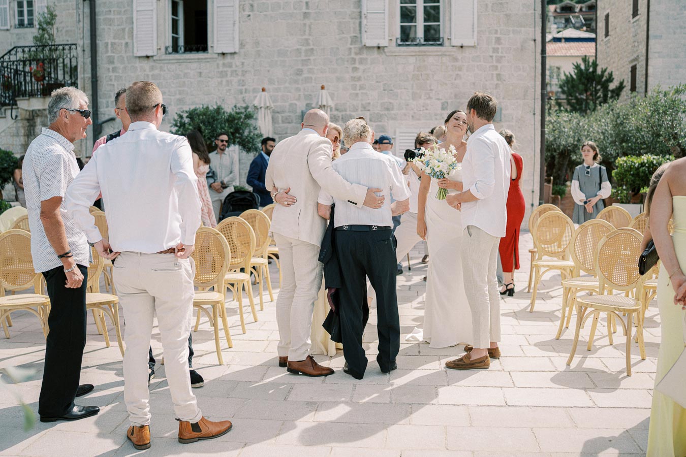 Wedding guests gathering outdoors in a sunny courtyard with stone buildings in the background. Some attendees are wearing formal attire and seated on wicker chairs, while others stand and chat. A person holds a bouquet, suggesting a wedding celebration.