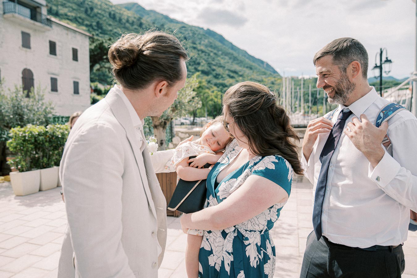 A family gathering outdoors, featuring a man in a beige suit, a woman in a floral blue dress holding a child, and a man in a white shirt with a tie smiling. Scenic background with greenery and hills.