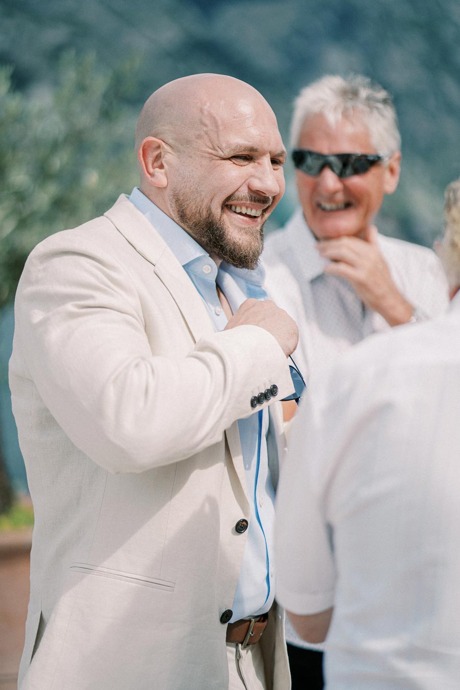 A smiling man in a light-colored suit adjusts his tie while standing outdoors, with blurred greenery in the background. Two other individuals are partially visible, engaging in conversation. The scene exudes a cheerful and relaxed atmosphere.