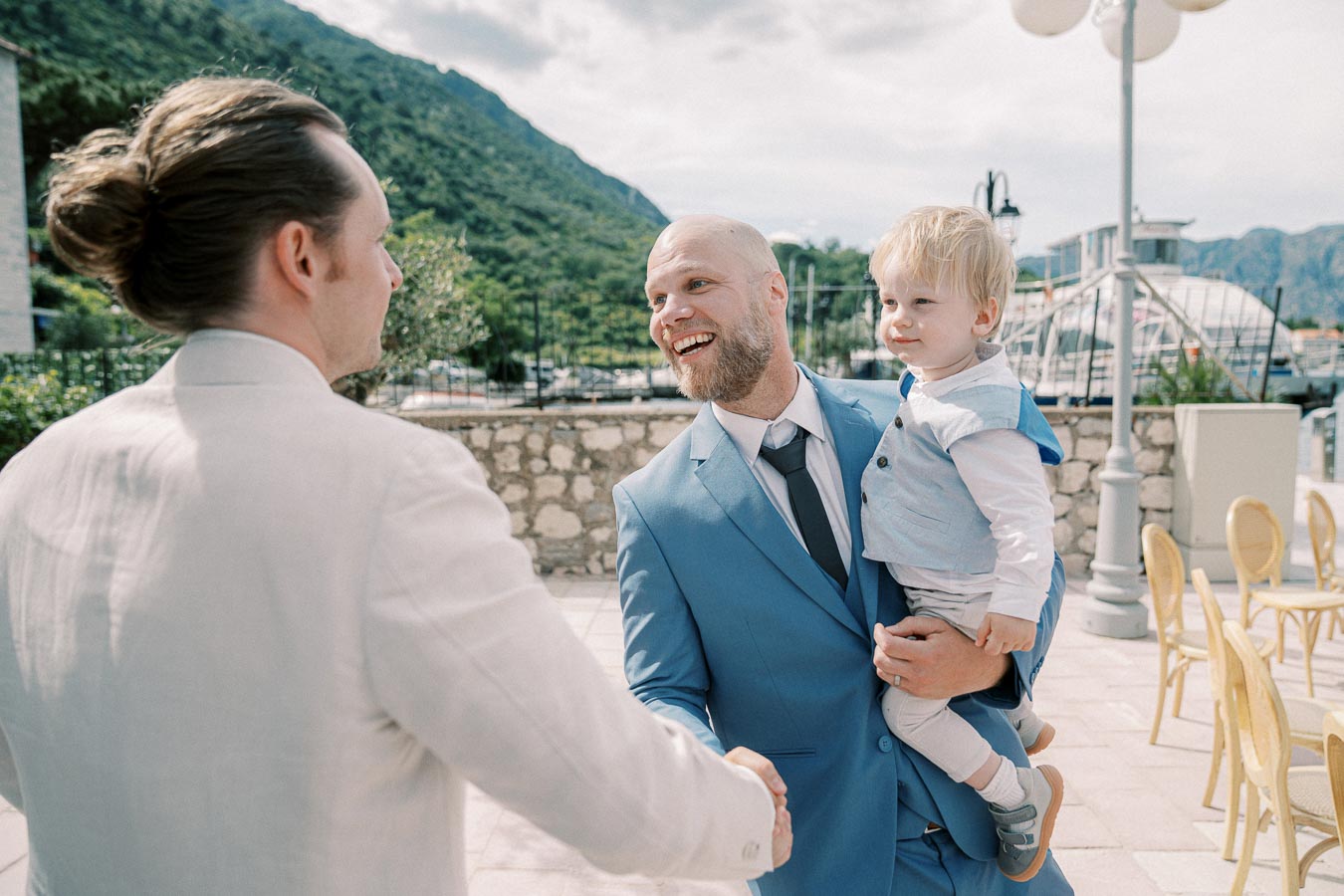 A man in a blue suit smiling while holding a young child, shaking hands with another man in a white suit, set against a scenic outdoor background of mountains and a marina.