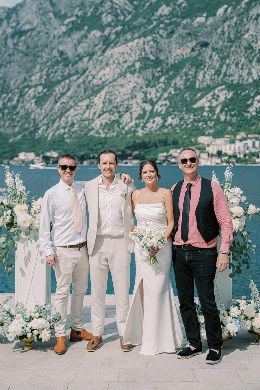 A wedding couple stands between two friends, all smiling, in front of a stunning mountain and lake backdrop, with decorative white floral arrangements on either side. The scene captures a sunny outdoor ceremony by the water.