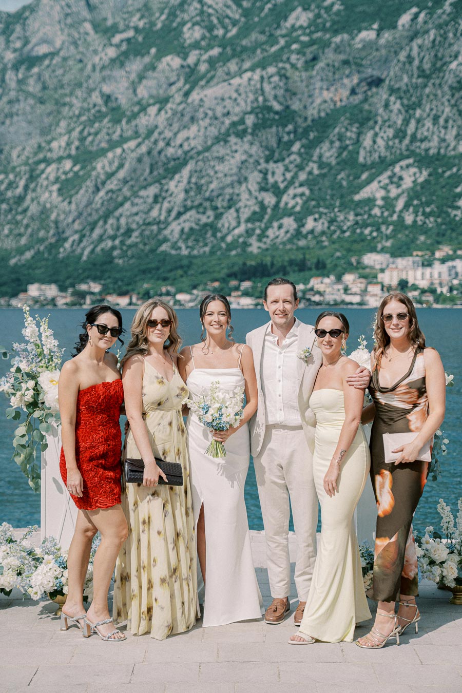 A group of six people dressed elegantly pose outdoors with a mountain and lake backdrop, capturing a scenic wedding or celebration moment.