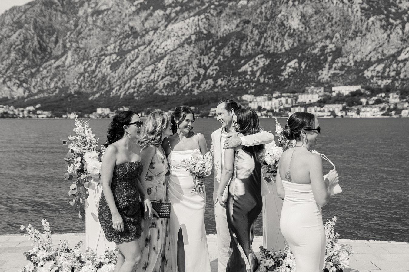 Bridal party celebrating at a lakeside wedding with stunning mountain backdrop, featuring elegantly dressed guests and floral arrangements. Black and white photography.