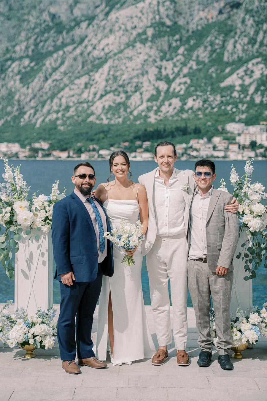A wedding photo featuring a bride and groom standing between two male guests, posing happily by a picturesque waterfront. Decorative floral arrangements and majestic mountains provide a stunning backdrop. The sky is clear, adding to the serene ambiance of the celebration.