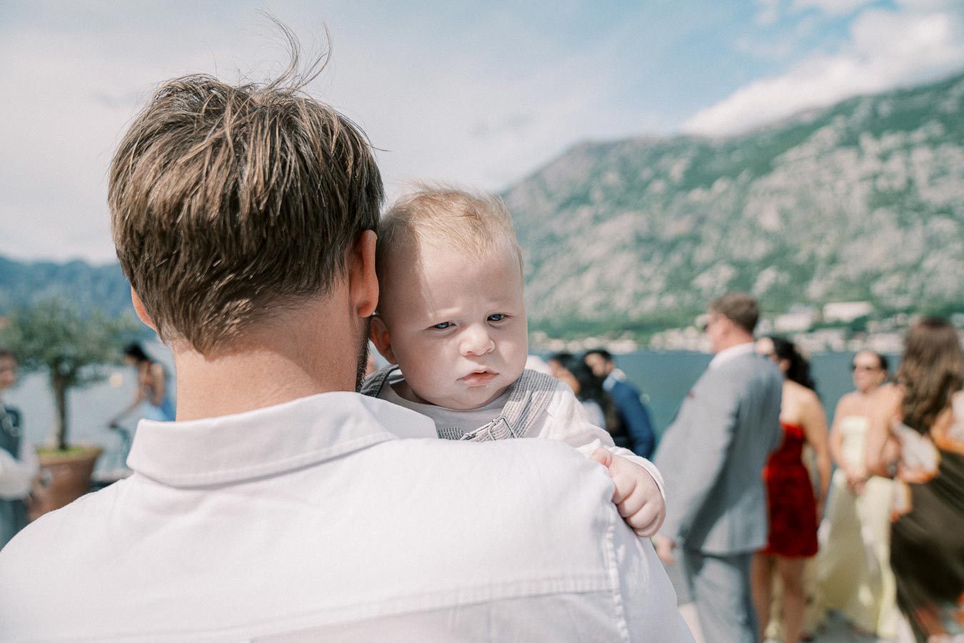 Father holding baby at an outdoor gathering near a scenic lake with mountains in the background.