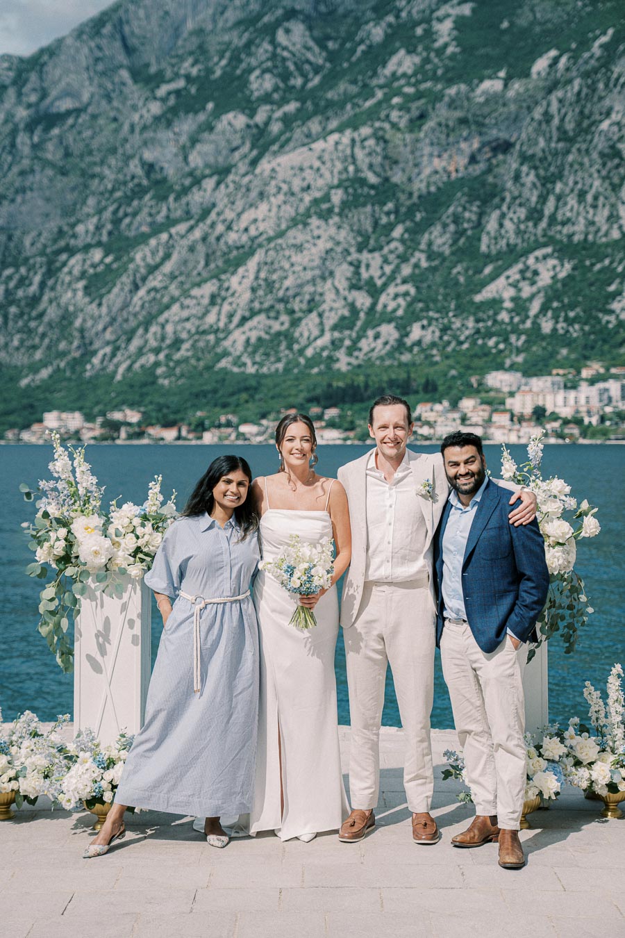 A group of four people standing happily in front of a scenic lake and mountain backdrop, with elegant floral arrangements, during a sunny outdoor wedding ceremony.