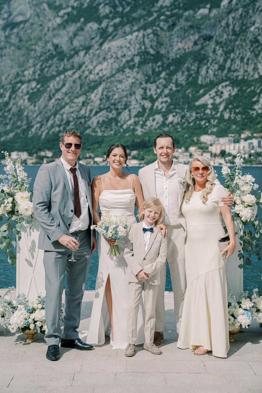 A happy wedding group poses outdoors with a scenic mountain and lake backdrop. The bride in a white dress holds a bouquet, surrounded by the groom and guests dressed in formal attire, including a child in a suit and bow tie. Beautiful floral arrangements add to the festive atmosphere.