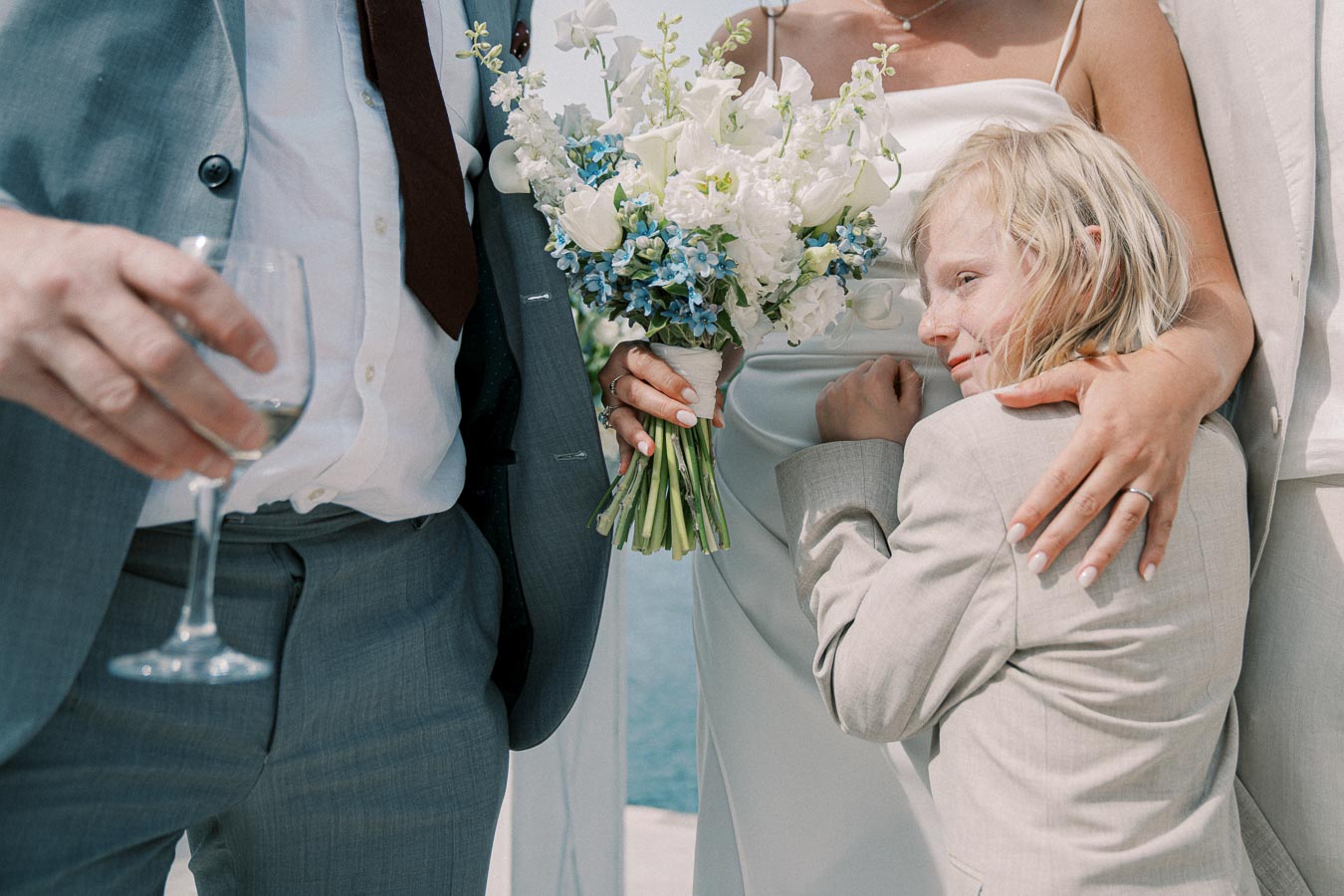A child in a gray suit affectionately hugs an adult holding a bouquet of blue and white flowers, with another adult holding a wine glass nearby, all dressed in formal attire, celebrating an outdoor occasion.