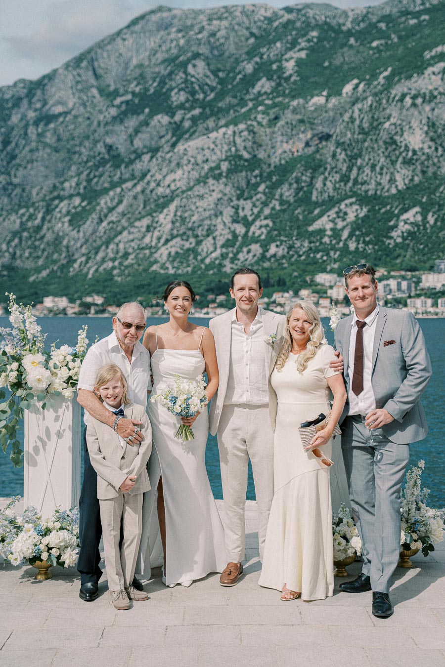 Elegant wedding party posing by scenic mountain lake backdrop with floral decorations.