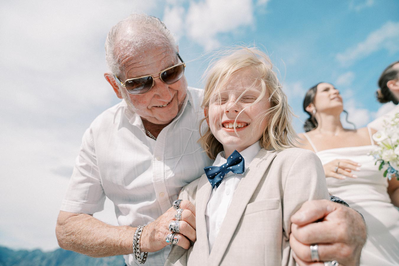Elderly man with sunglasses joyfully holding a smiling child's shoulders at an outdoor event, both well-dressed with a clear blue sky in the background.