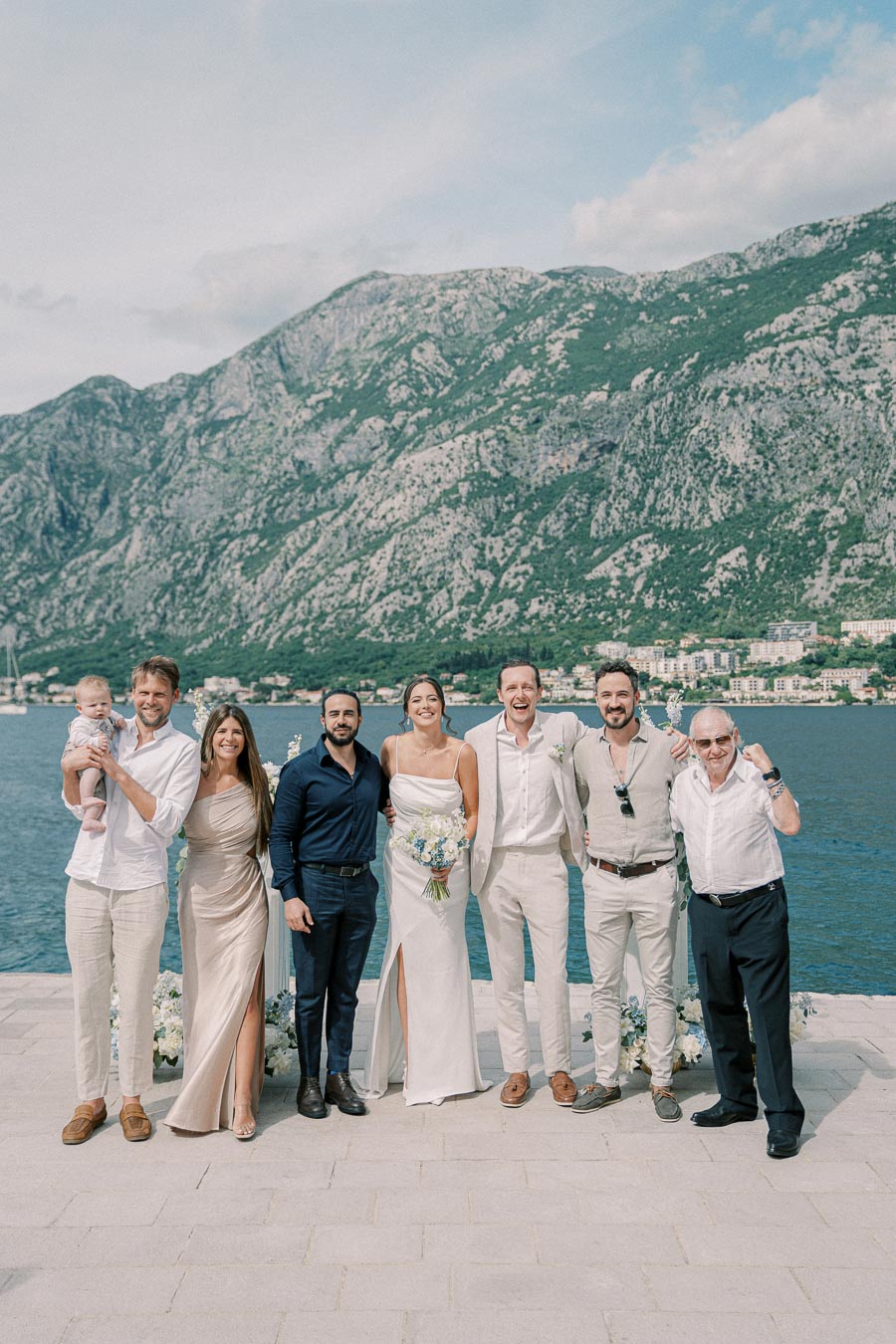 A group of people, including a bride in a white dress holding a bouquet, posing at a wedding with a picturesque mountain and lake backdrop on a sunny day.