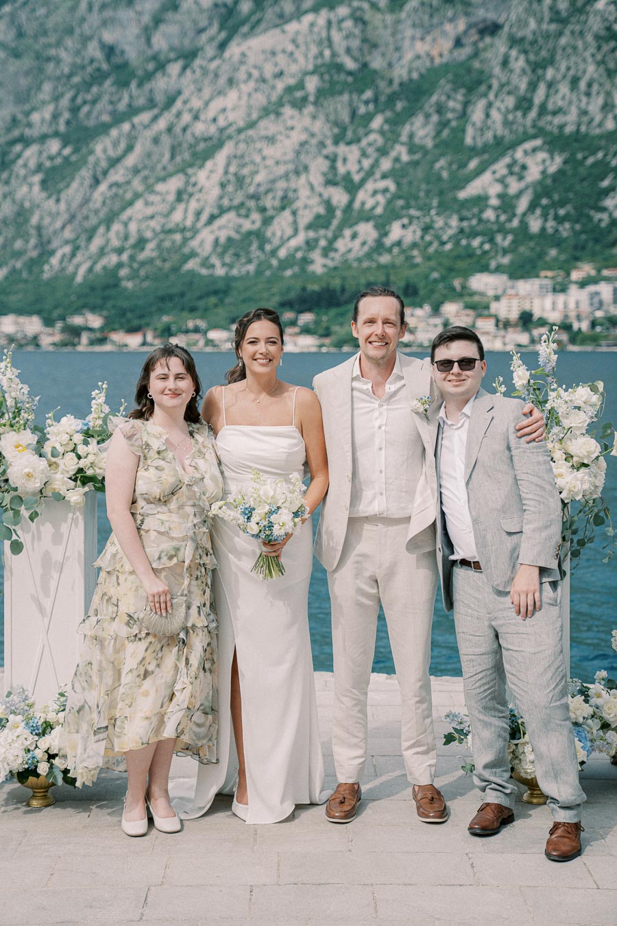 A group of four people dressed in formal attire poses happily at a waterfront wedding ceremony, surrounded by floral arrangements, with a scenic view of mountains and the sea in the background.