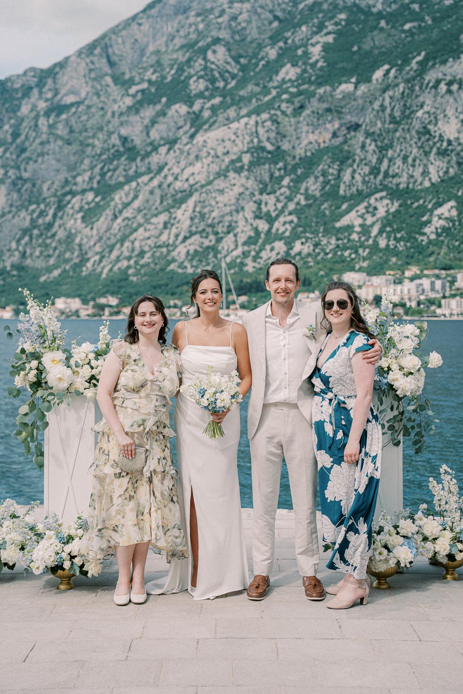 Wedding party posing by scenic waterfront with mountains, featuring floral arrangements, elegant attire, and a joyful atmosphere.