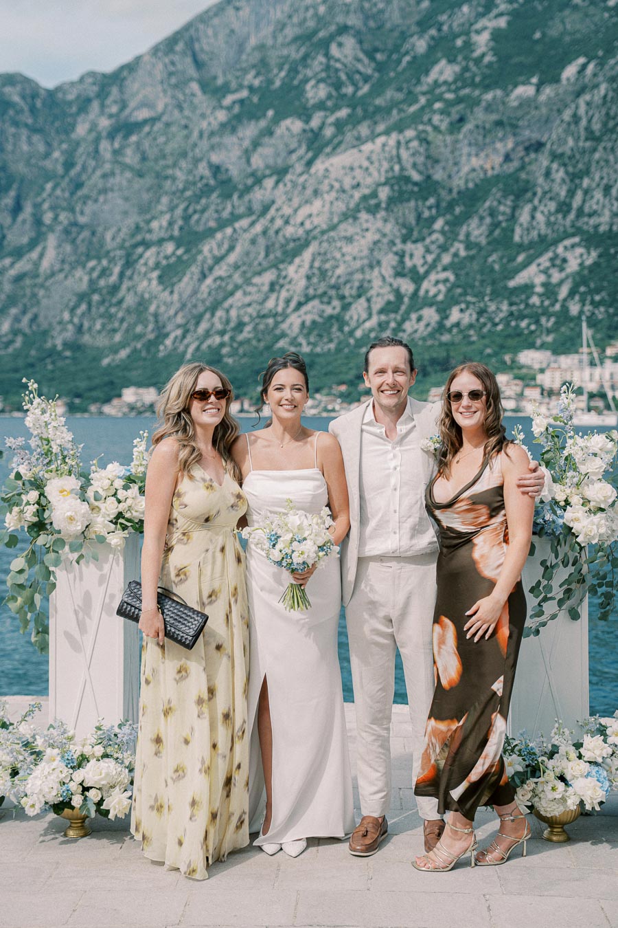 Four people posing at an outdoor wedding ceremony by the sea, with lush mountains in the background. The bride in a white dress holds a bouquet, while the groom and two guests stand beside her, all smiling amid floral arrangements.