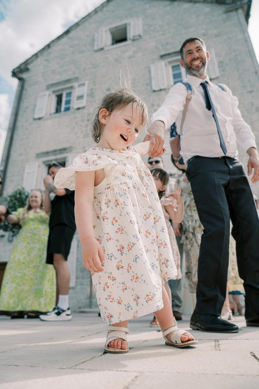 A joyful child in a floral dress smiling and holding hands with an adult in professional attire, standing outside a stone building on a sunny day, with a group of people in the background.