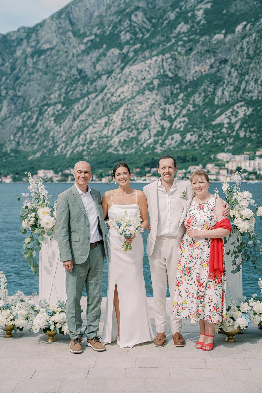 A wedding party posing in front of a scenic mountain and lake backdrop, featuring the bride holding a bouquet, the groom, and two other guests, adorned with floral arrangements and dressed in formal attire.