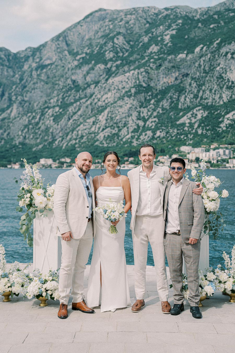 Wedding party posing outdoors by a scenic lake with mountains in the background. The couple is surrounded by two men, all dressed in light-colored suits. Floral arrangements add elegance to the ceremony setting.