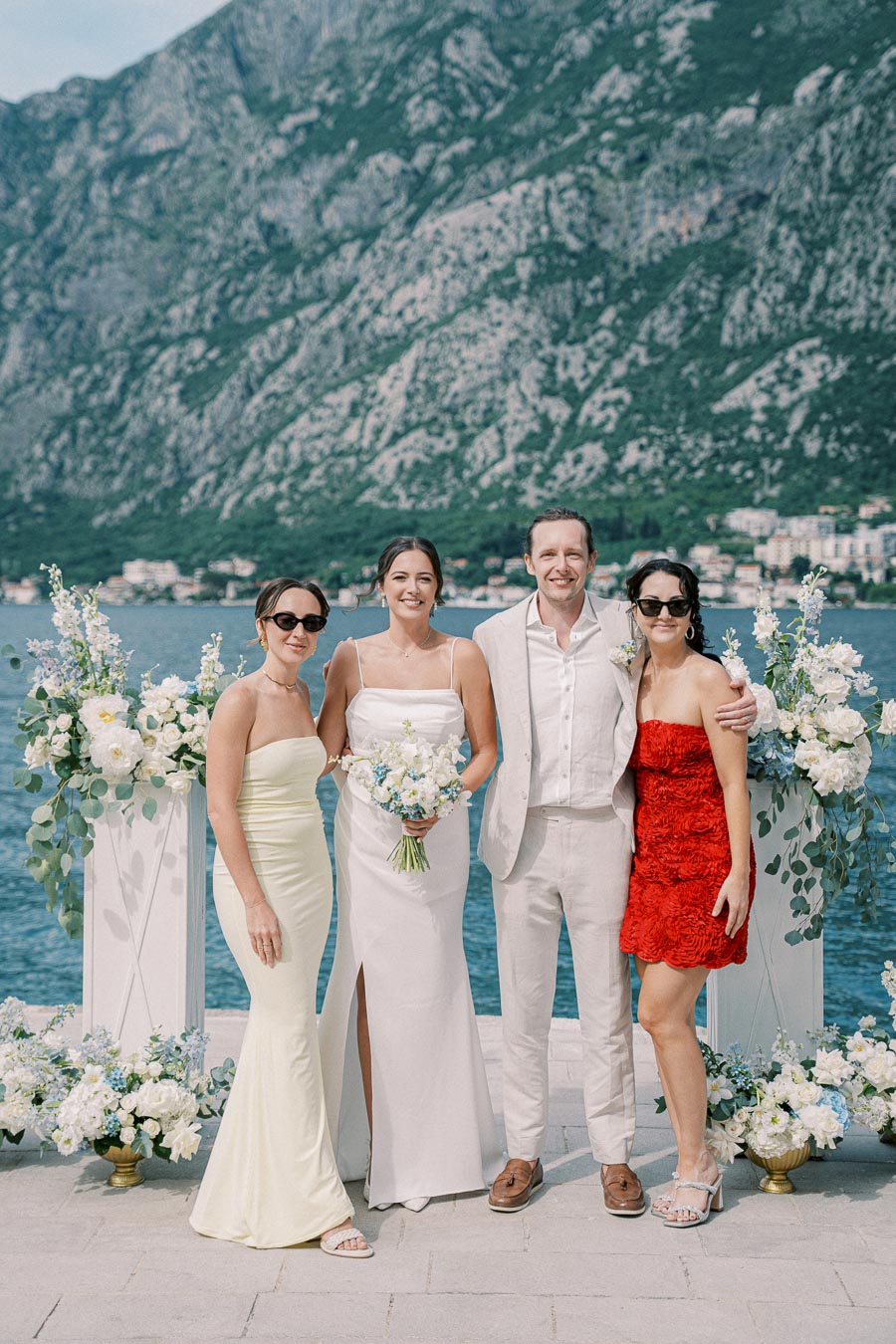 A group of four people dressed in formal attire, standing together at an outdoor wedding ceremony by the water, with mountainous scenery in the background and floral decorations.