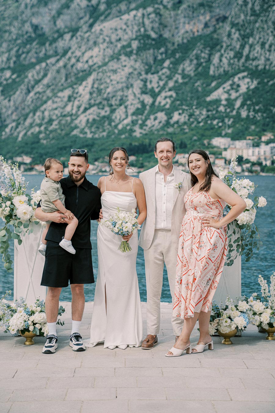 Group photo at a scenic outdoor wedding with mountainous backdrop, featuring a bride in a white dress holding a bouquet, a groom in a light suit, and three guests including a man holding a toddler.