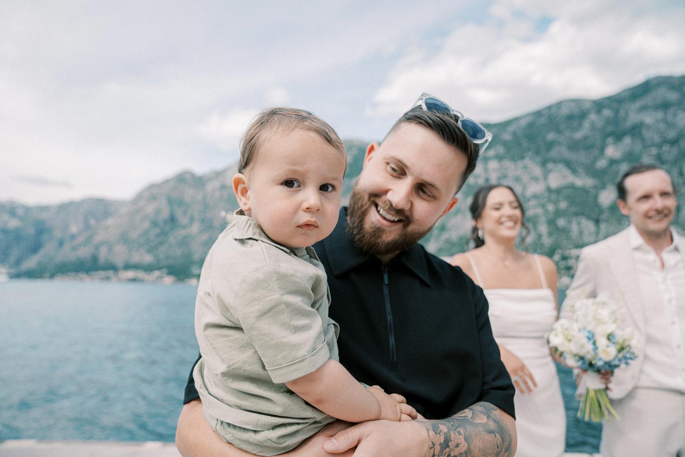 A joyful man holding a young child with a scenic mountain and lake backdrop, while a smiling bride and groom stand in the background at an outdoor wedding celebration.