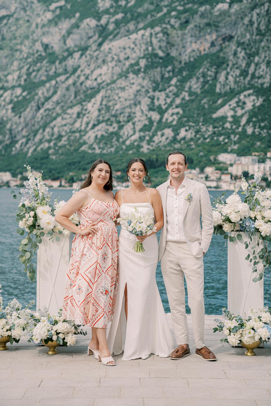Elegant wedding party posing by a scenic waterfront with mountainous backdrop, featuring a bride in a white dress holding a bouquet, a groom in a light suit, and a guest in a patterned dress, surrounded by floral arrangements.