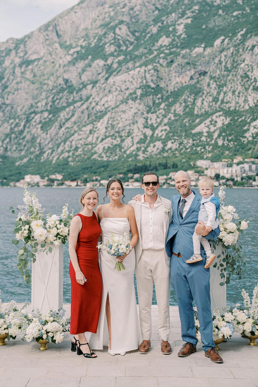 A wedding party poses with scenic mountain and lake backdrop, featuring a bride in a white dress holding a bouquet, surrounded by elegantly dressed guests in a vibrant outdoor setting.