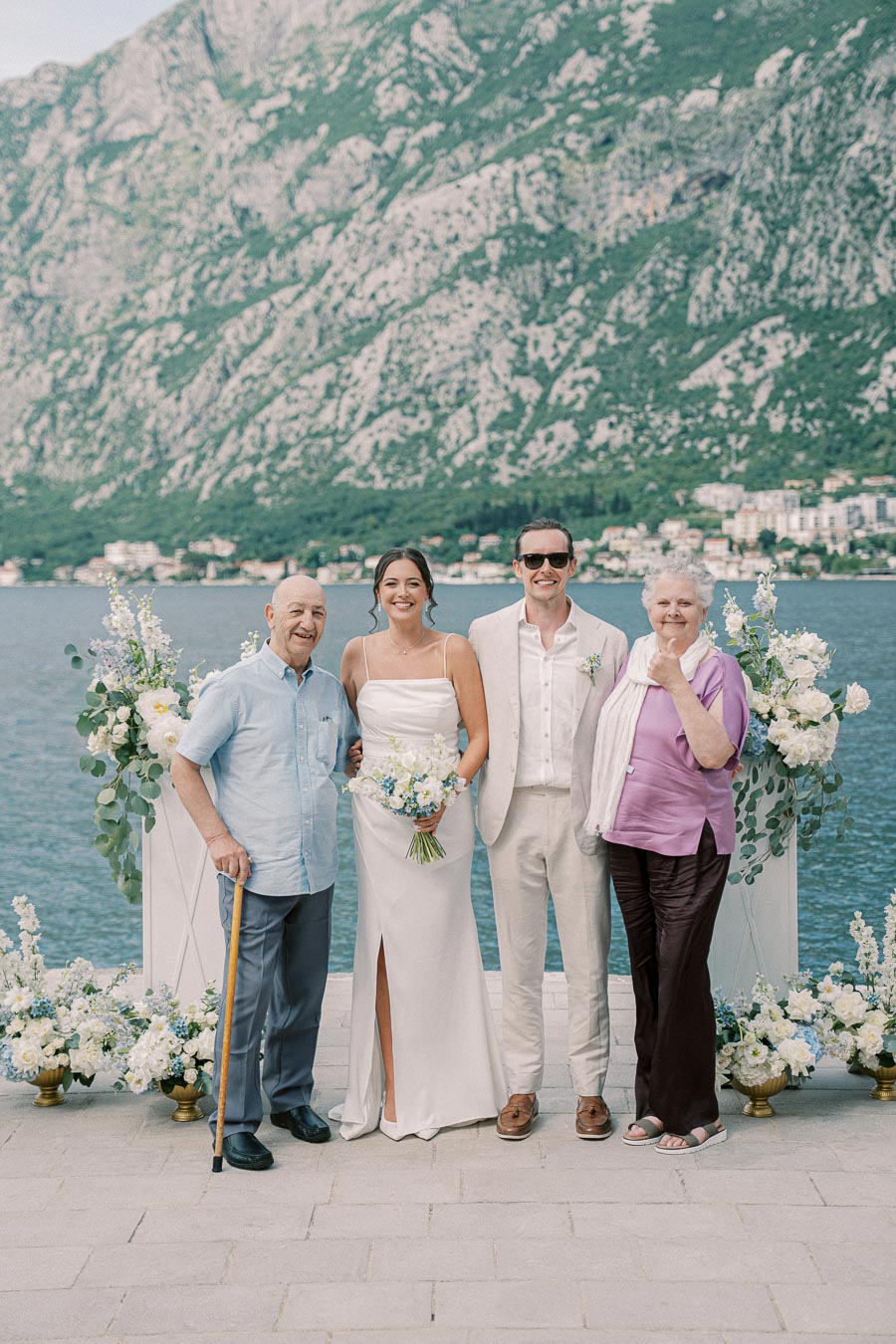 Wedding couple posing with family on a scenic waterfront, with decorative floral arrangements and majestic mountains in the background.