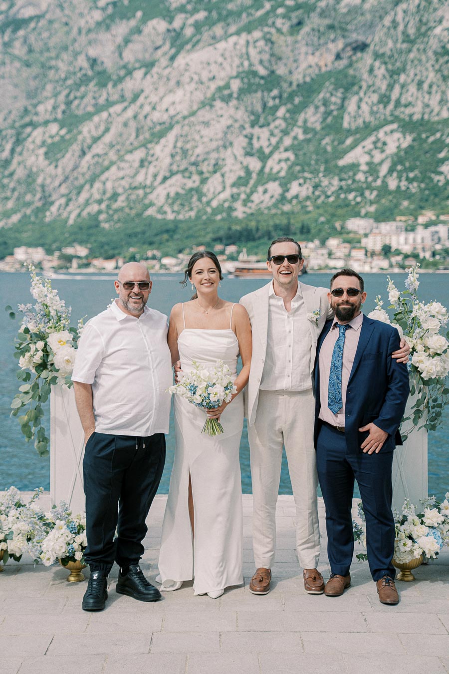 A bride and groom, along with two guests, posing together with a picturesque mountain and lake backdrop, featuring elegant floral arrangements on a sunny day.