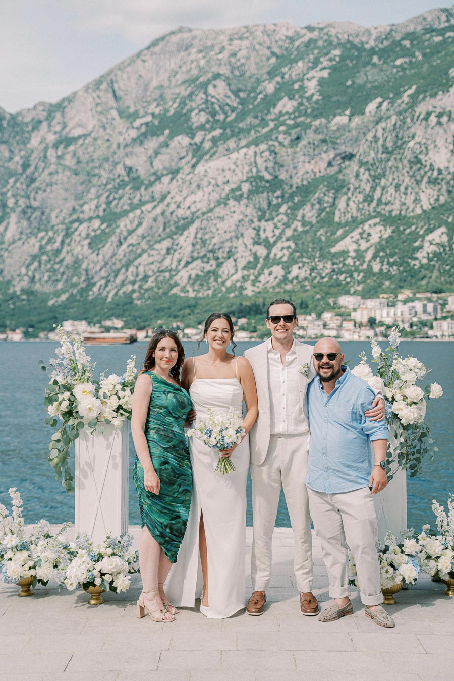 Wedding party posing by a scenic waterfront with mountain backdrop, facing the camera against elegant floral arrangements.