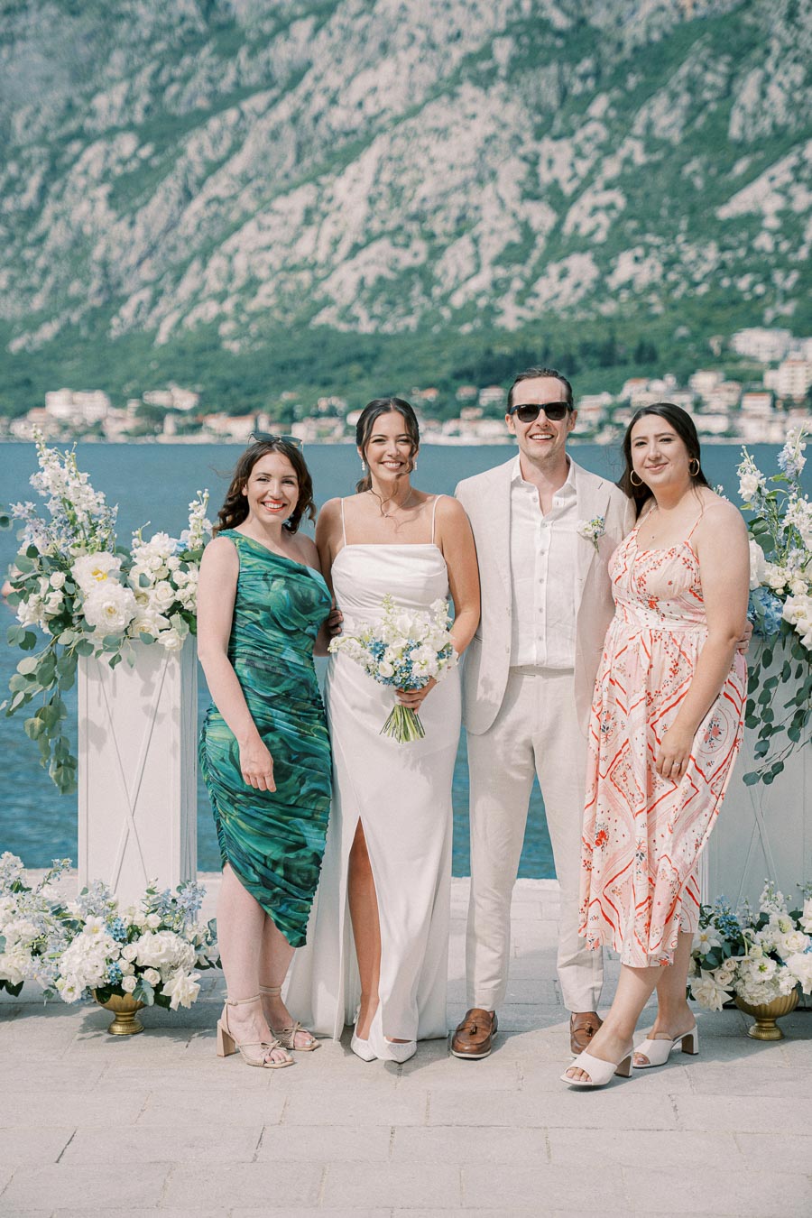 A group of four people posing together at a scenic outdoor wedding by the sea, with a mountainous backdrop. The bride is holding a bouquet, standing between two women in colorful dresses and a man in a light suit. Floral arrangements decorate the setting, adding to the elegant, festive atmosphere.