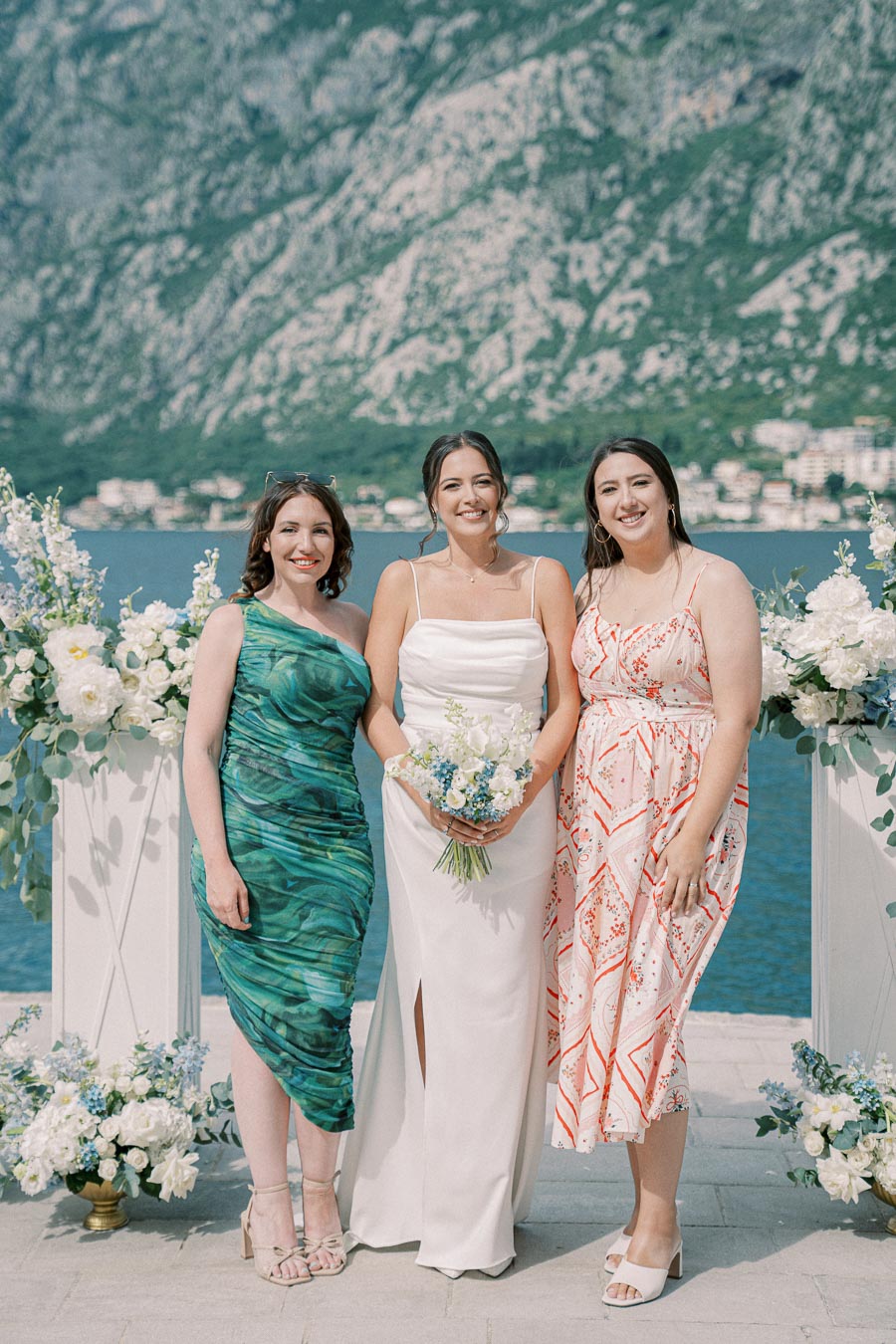 Three women posing together at a waterfront wedding, surrounded by floral arrangements, with a scenic mountain backdrop. The bride holds a bouquet of white and blue flowers.