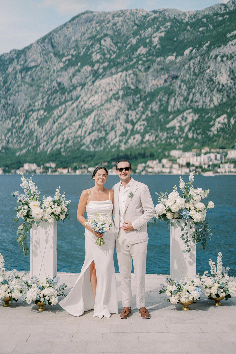 Bride and groom smiling at their outdoor wedding ceremony by the sea, surrounded by elegant floral arrangements and a mountainous backdrop.