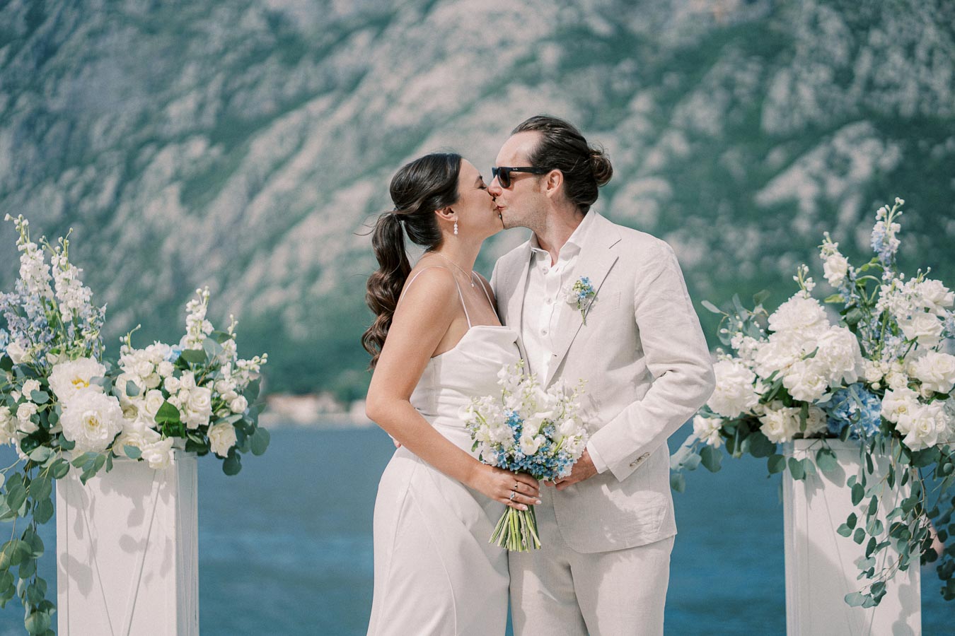 A couple shares a kiss in a scenic outdoor wedding ceremony with floral arrangements, set against a stunning mountainous backdrop and a lake.