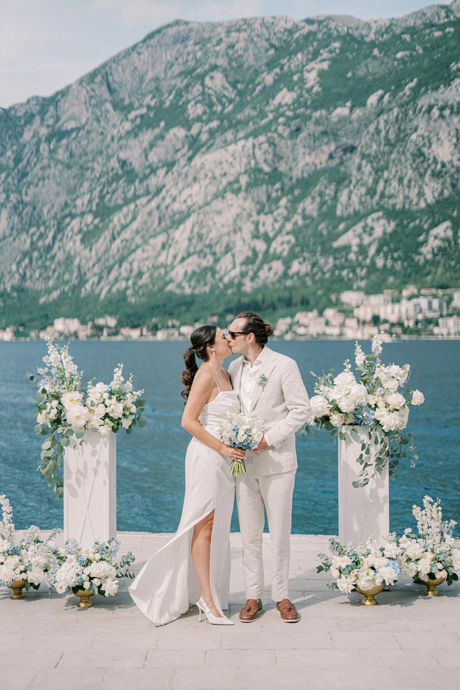A bride and groom share a romantic kiss during an outdoor wedding ceremony by a picturesque waterfront, surrounded by elegant floral arrangements, with majestic mountains in the background.