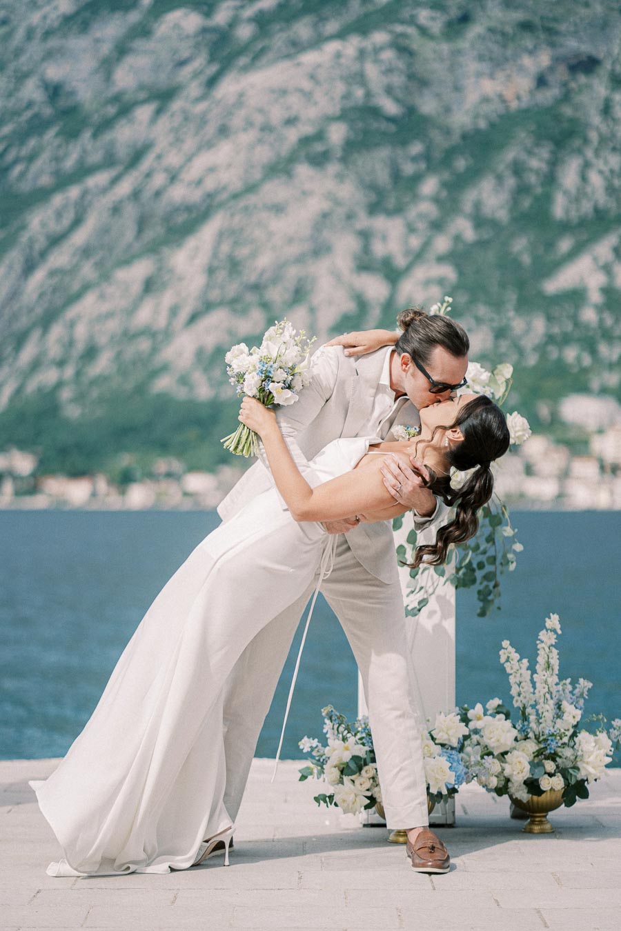 A couple kissing passionately during an outdoor wedding ceremony by the sea, with mountains in the background and elegant floral arrangements, both wearing white attire.