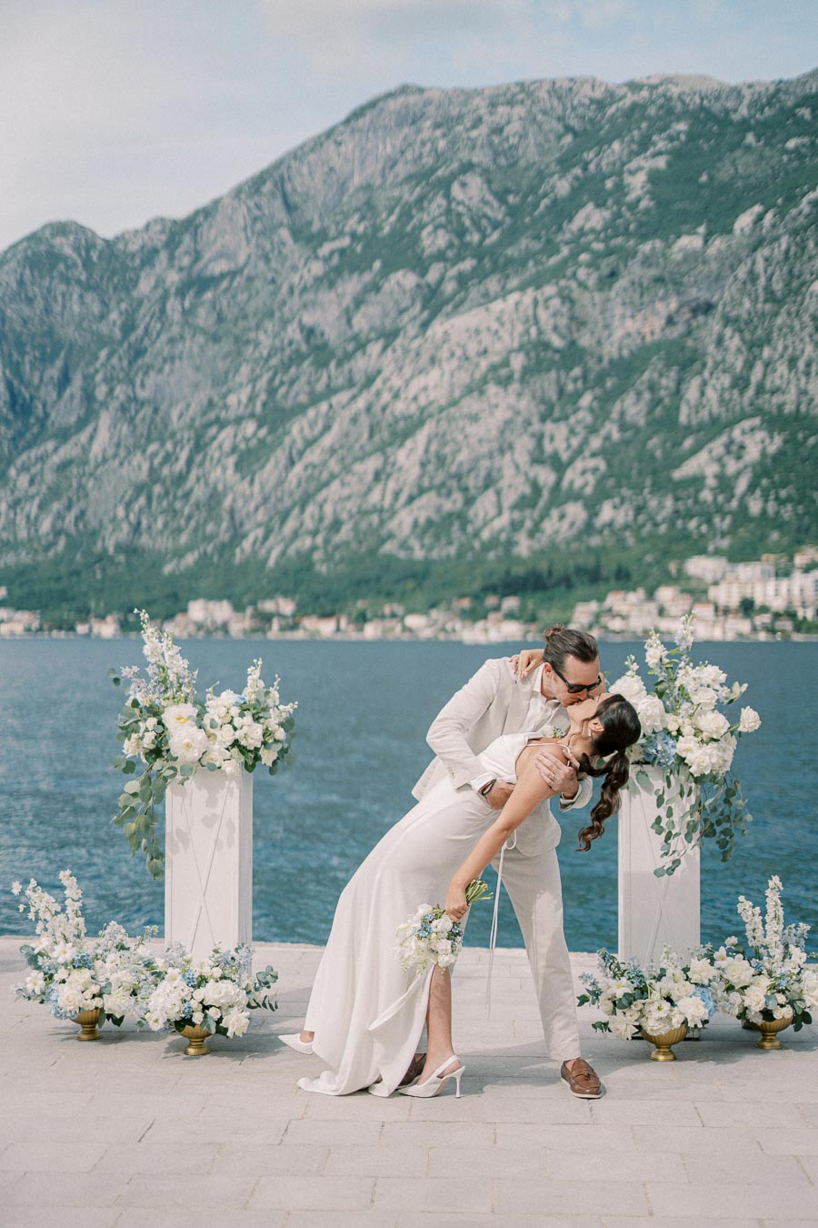 A bride and groom share a romantic kiss by the seaside with a backdrop of picturesque mountains and elegant floral arrangements.