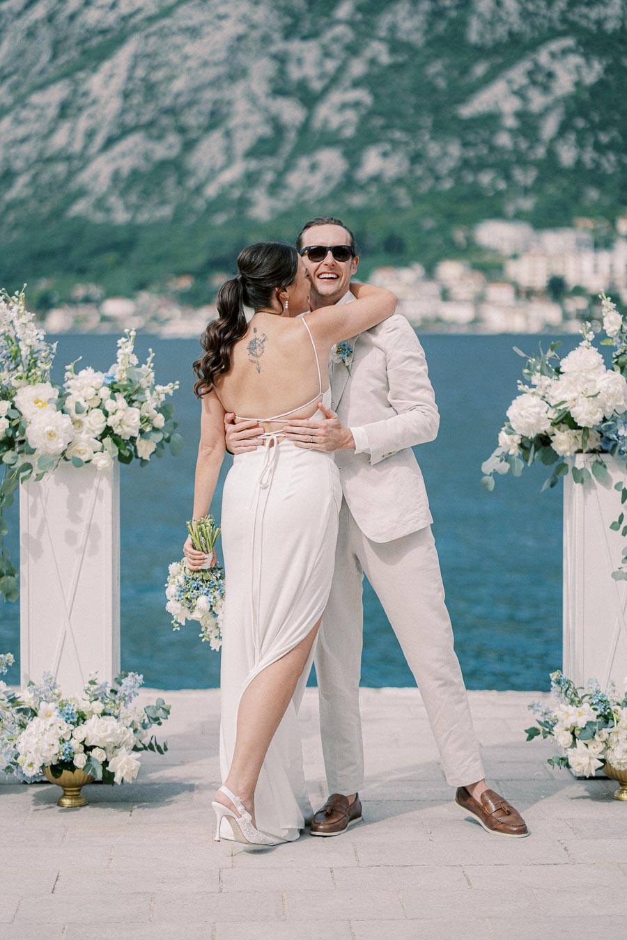 A joyful couple embracing during their outdoor wedding ceremony by the sea, surrounded by elegant white and blue floral arrangements, with a scenic mountainous backdrop.