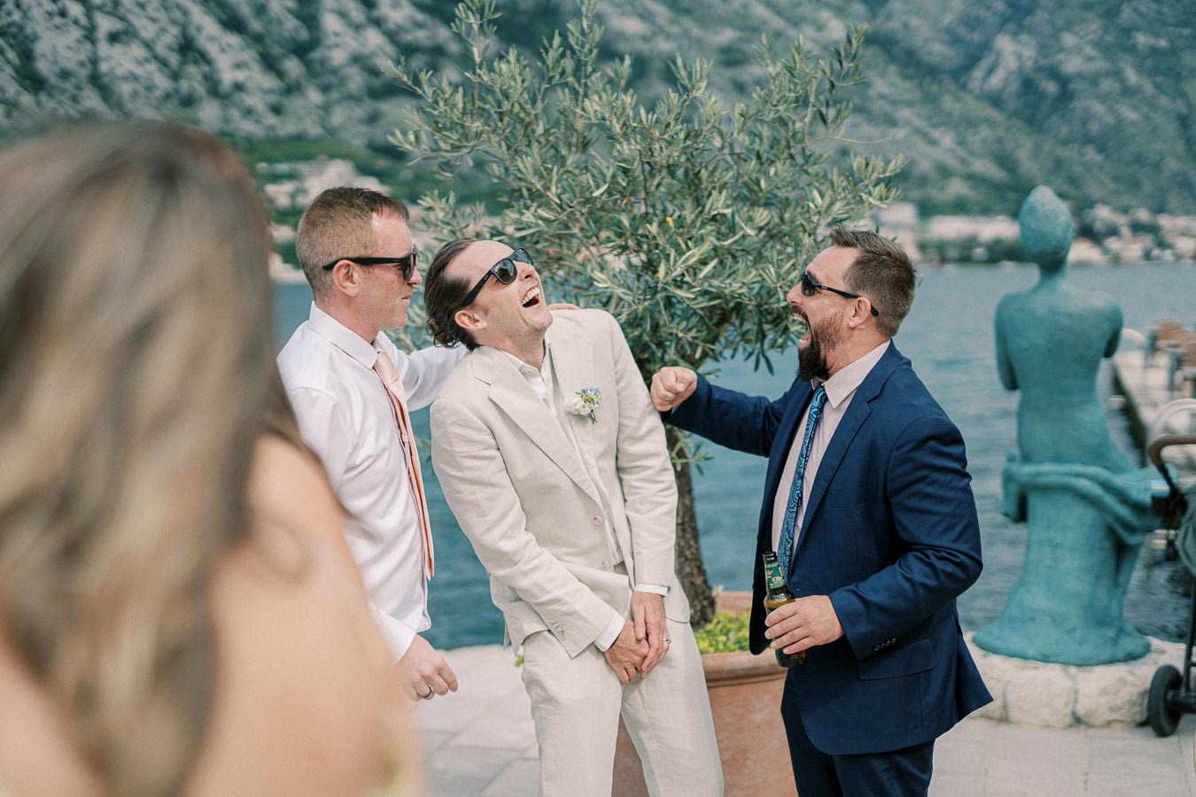 Three men in suits laughing together at a waterfront wedding reception, with a scenic mountain view in the background.