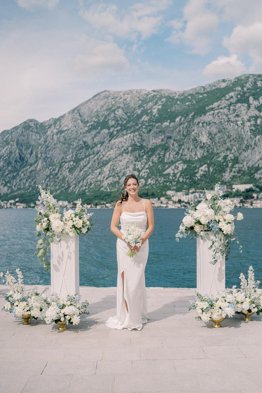 Bride in elegant white wedding dress standing by a scenic waterfront with mountains in the background, adorned with floral arrangements on a sunny day.