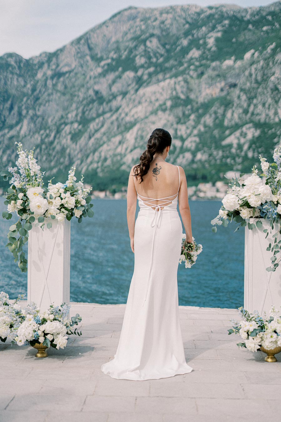 Bride in elegant white dress with back tattoo stands between lush floral arrangements, overlooking a serene lake with majestic mountains in the background.