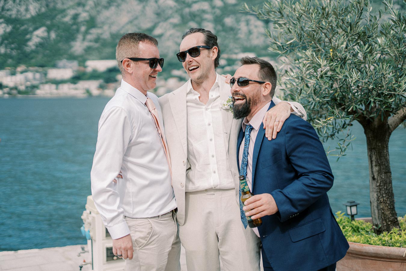 Three men in formal attire, smiling and embracing, stand near a scenic waterfront with mountains in the background. They are enjoying a celebration, possibly at a wedding or outdoor event, with one holding a bottled drink. The atmosphere is joyous and relaxed.