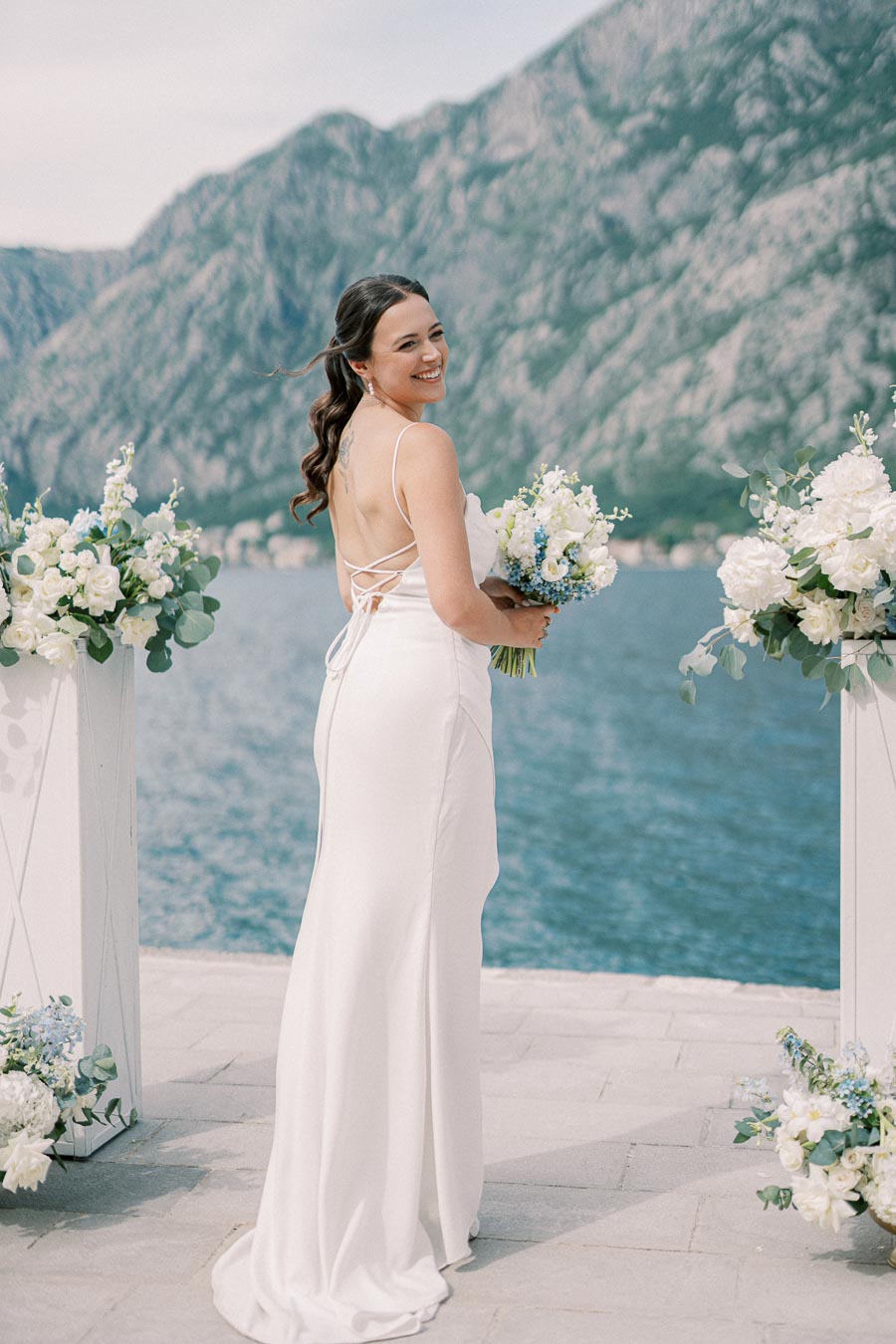 Bride in elegant white dress holding a bouquet, smiling at an outdoor wedding ceremony by a scenic lake with mountain views.