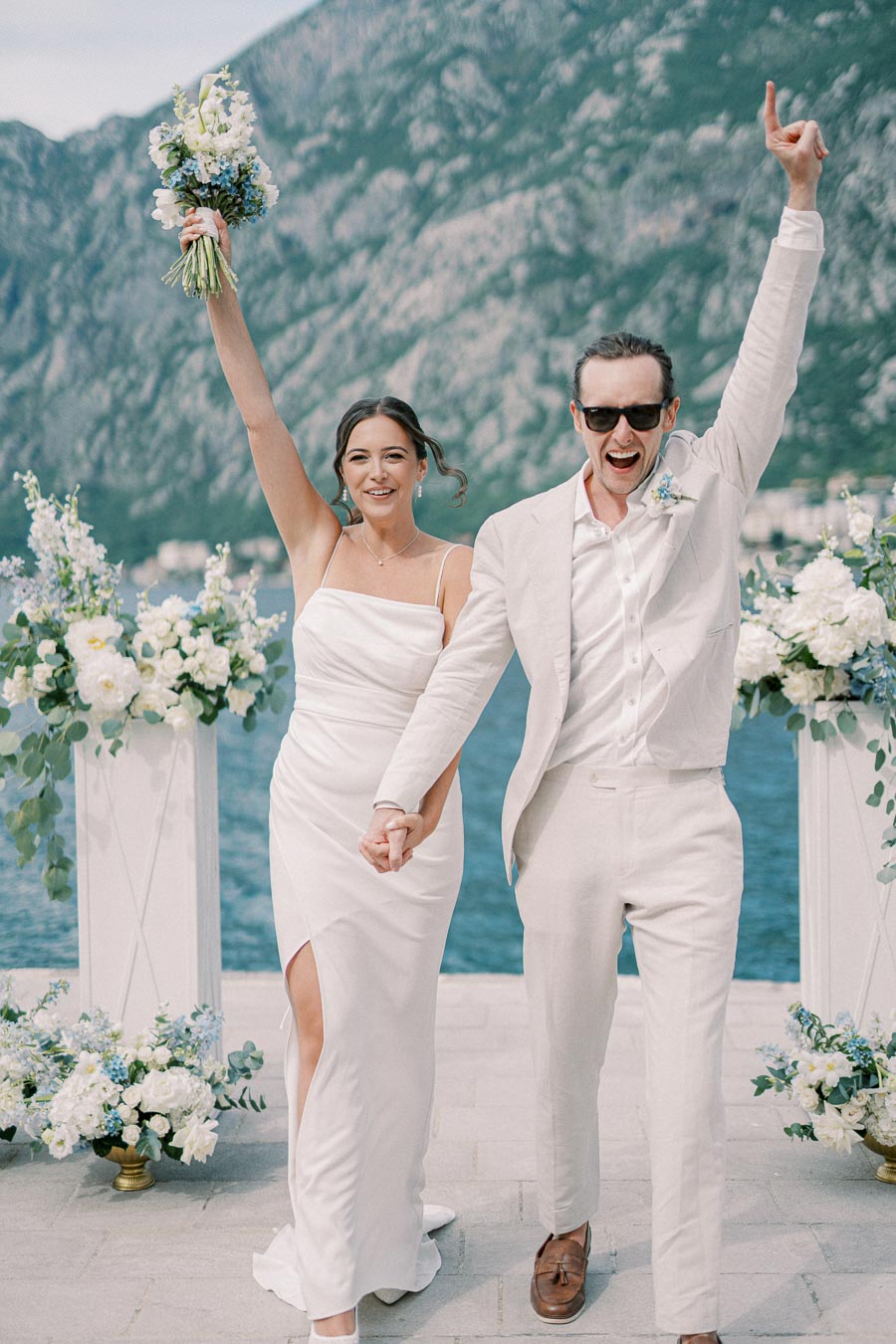 A joyful couple celebrates their wedding by a picturesque lakeside backdrop, adorned with elegant floral arrangements, under a clear sky, with a mountainous landscape in the background.