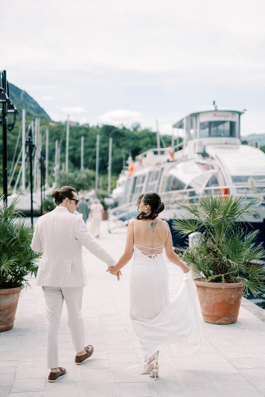A couple in elegant attire walks hand in hand along a marina lined with yachts, surrounded by palm plants, with scenic mountains in the background.