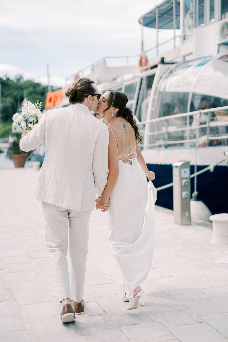 A newlywed couple in elegant white attire shares a romantic kiss by a marina, holding hands and a bridal bouquet, with a yacht in the background.
