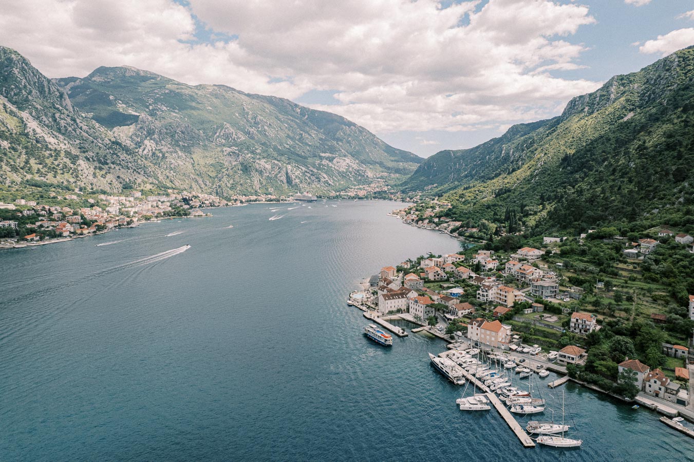 Aerial view of scenic coastal town with marina and boats in Montenegro, nestled between dramatic green mountains and blue sea under a cloudy sky.