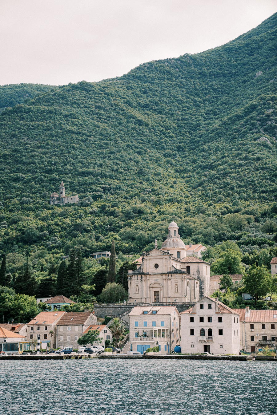 A picturesque seaside village nestled at the base of lush green hills in Montenegro, featuring historic architecture with a prominent dome structure, surrounded by traditional stone houses and a serene waterfront.