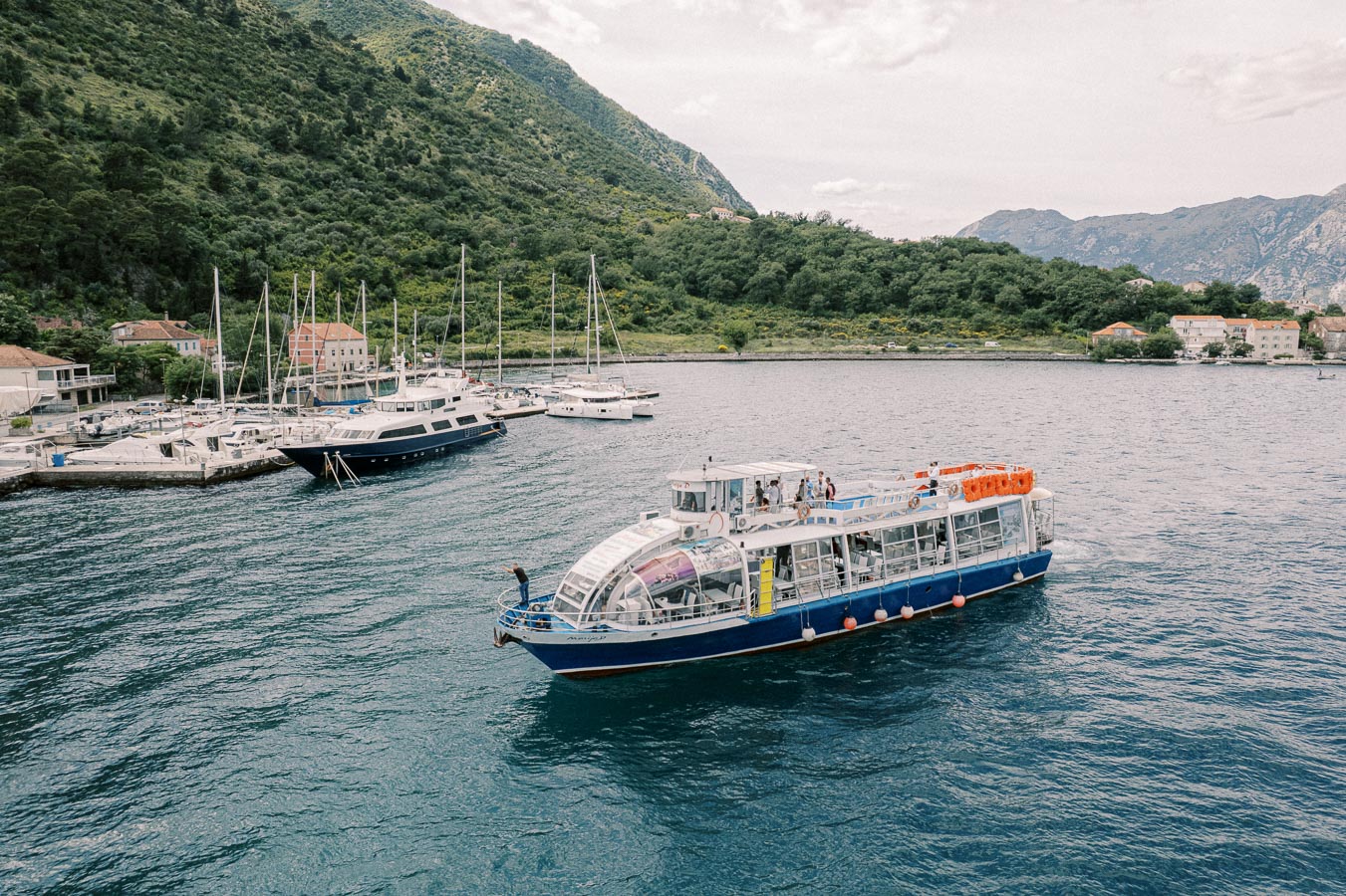A scenic view of a passenger boat sailing near a marina with several yachts docked, surrounded by lush green hills and quaint buildings, under a cloudy sky. Perfect for promoting coastal tourism and maritime activities.