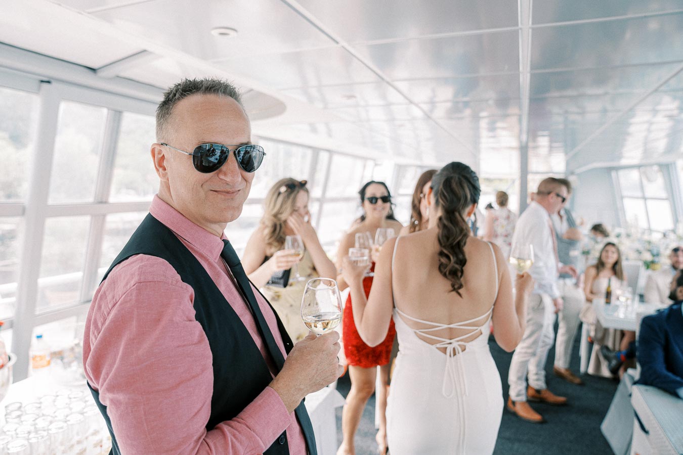 A stylish man in sunglasses holds a glass of wine at a lively indoor party, surrounded by well-dressed guests mingling and enjoying drinks.
